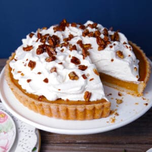 Pumpkin pie tart with lots of whipped cream on top as well as candied pecans on a white cake stand, blue background.