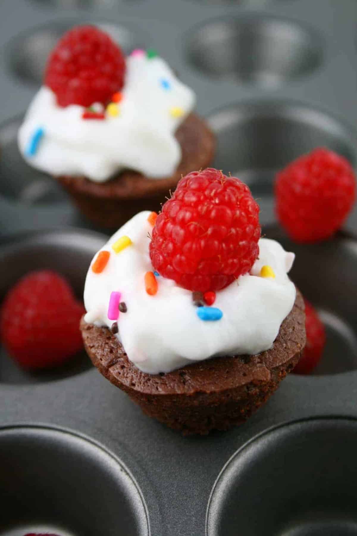 close up of two low calorie chocolate mini muffins display on a muffin pan, topped with whipped cream, multi colored sprinkles and raspberries