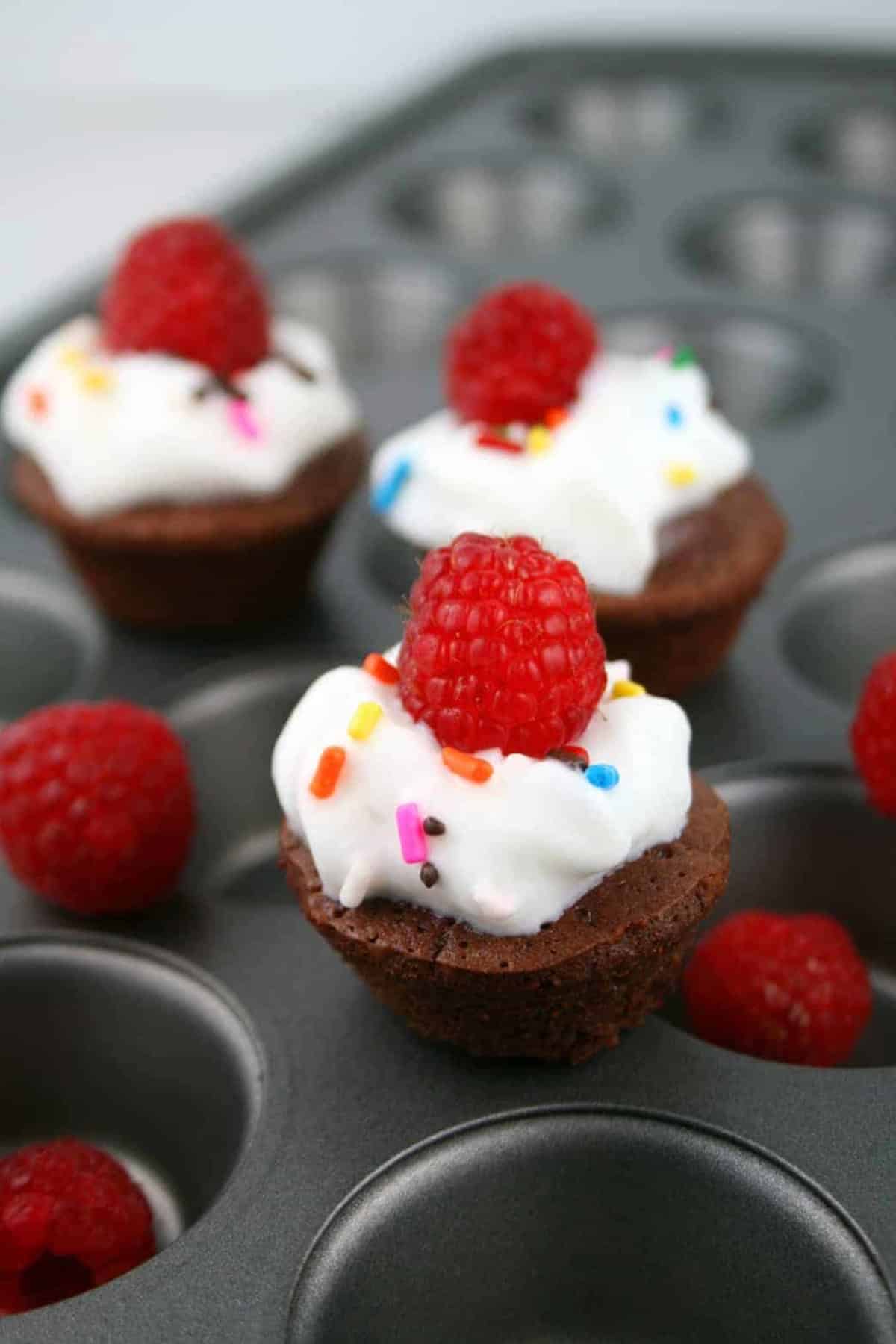 low calorie chocolate mini muffins display on a muffin pan, topped with whipped cream, multi colored sprinkles and raspberries.