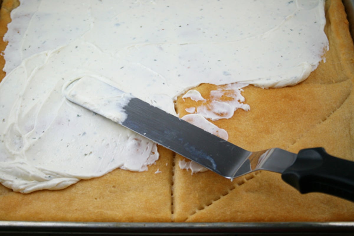 Spreading cream cheese Ranch onto cooled pizza crust.
