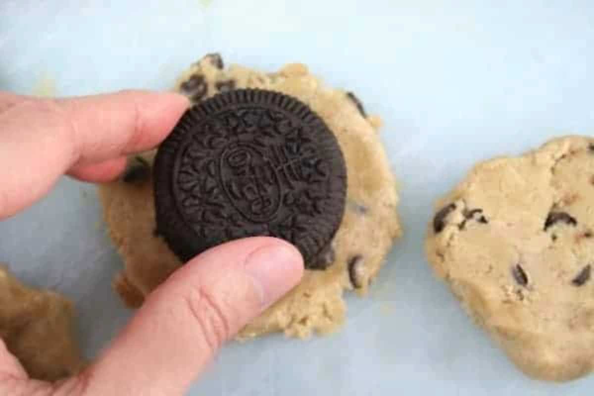 fingers holding an Oreo over chocolate chip cookie dough. 