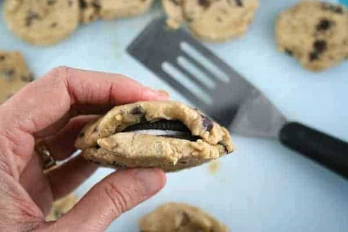 hand holding cookie showing inside pre baked Oreo stuffed chocolate chip cookies and metal spatula on table. 