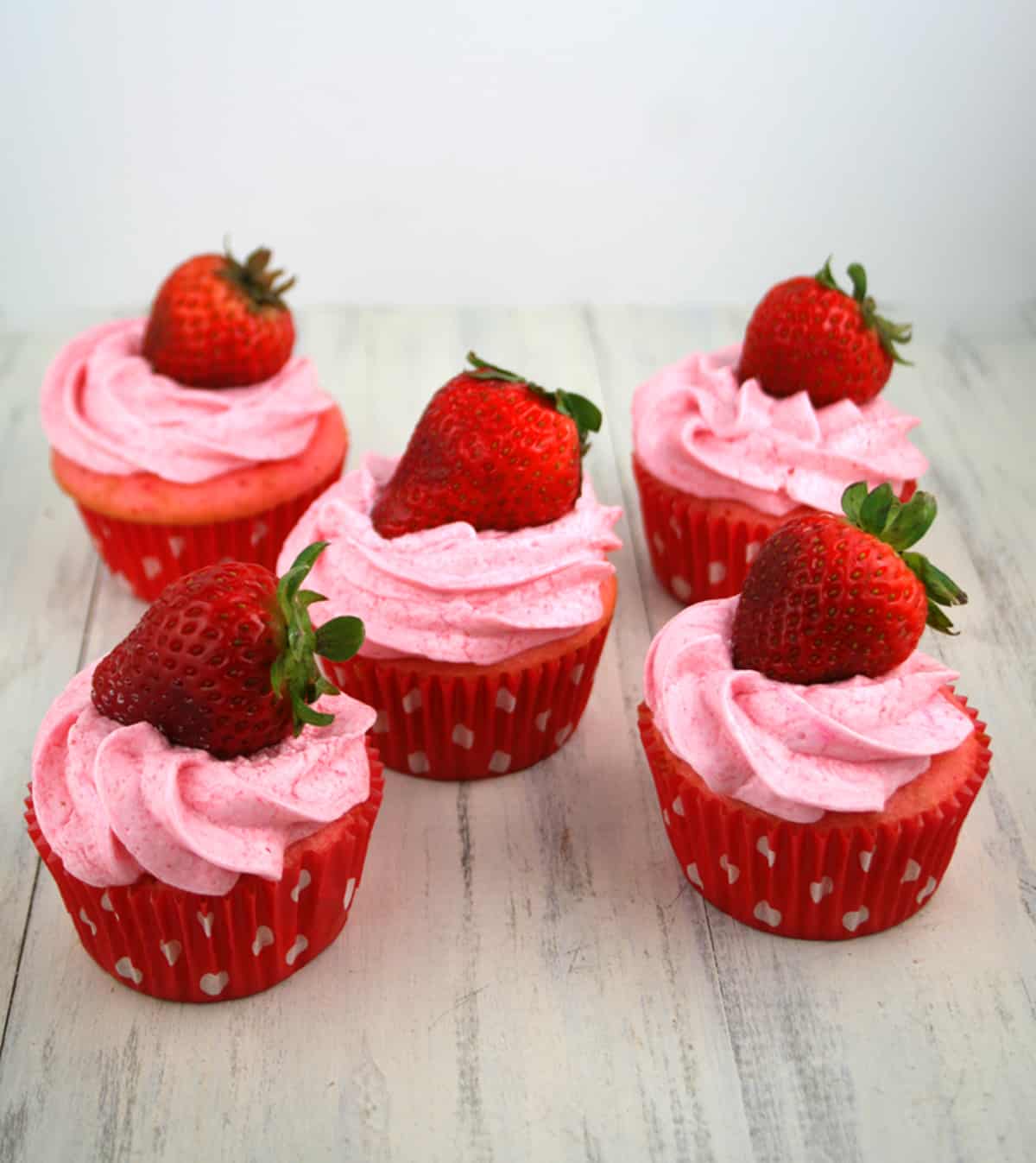 Five strawberry cupcakes in red cupcake liners with white polka dots and fresh strawberry on top. 