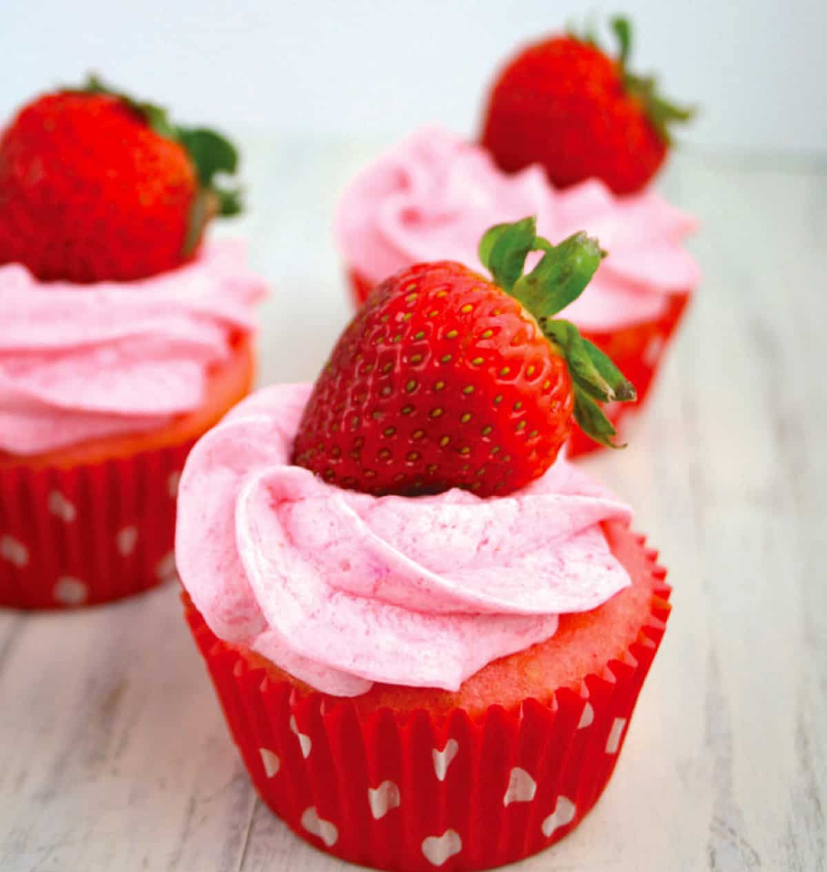 Three pink strawberry cake cupcakes with pink frosting and strawberry on top.