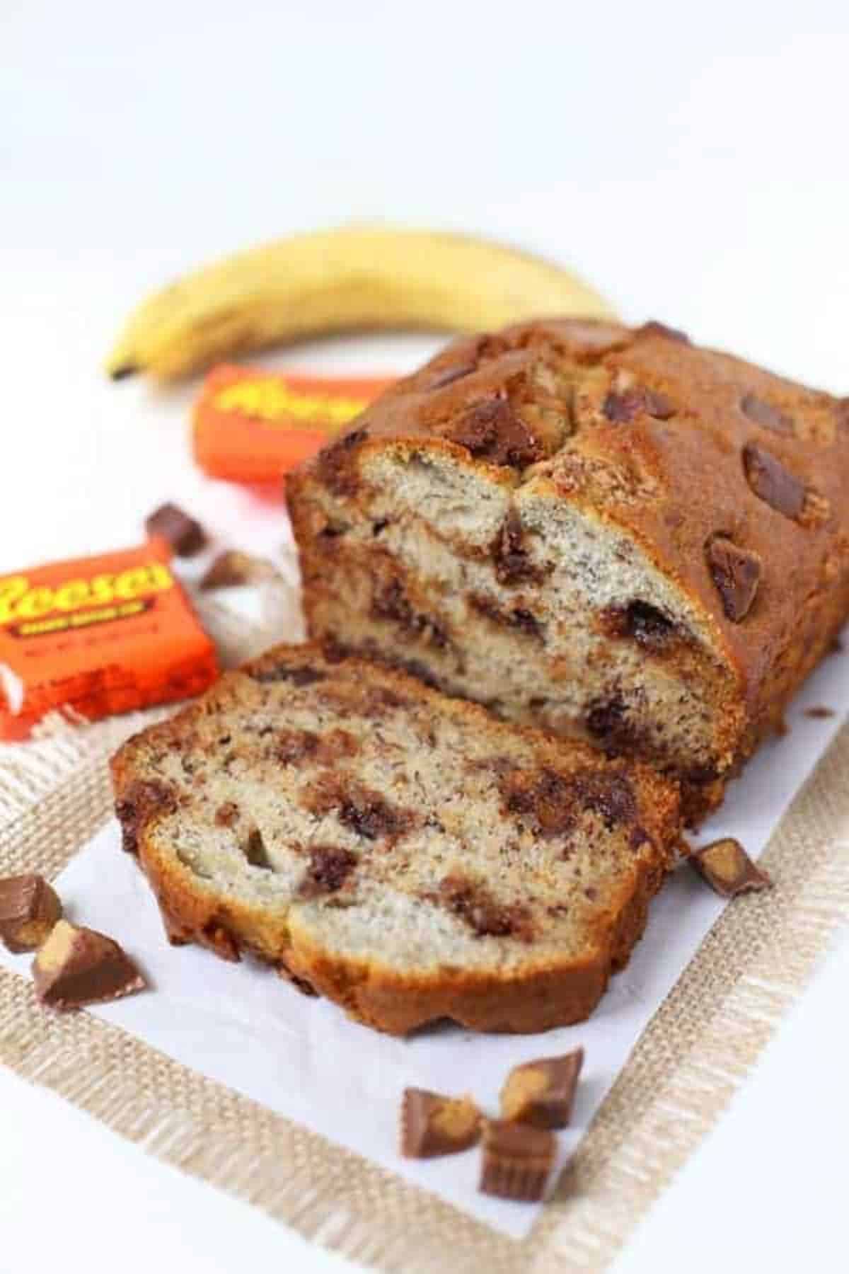Reeses Peanut Butter Cup Banana Bread with slice cut and chopped Reeses cups surrounding it. Banana and Reeses in packages in top left corner. 