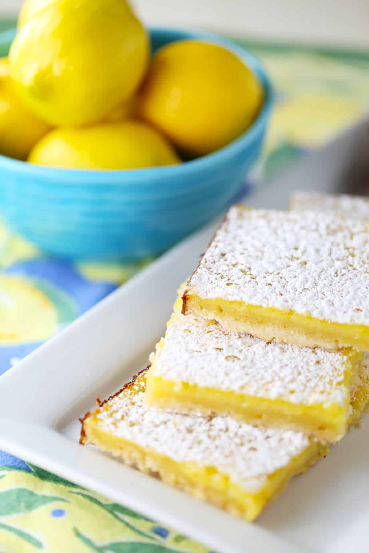 A stack of 3 lemon bars on a square white plate, with a blue bowl of lemons in the background.