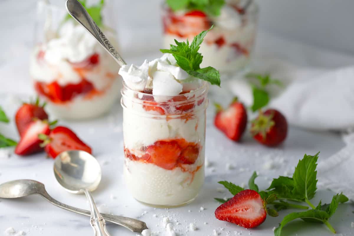 Three finished jars of the British dessert with cut strawberries on the counter next to the jars.