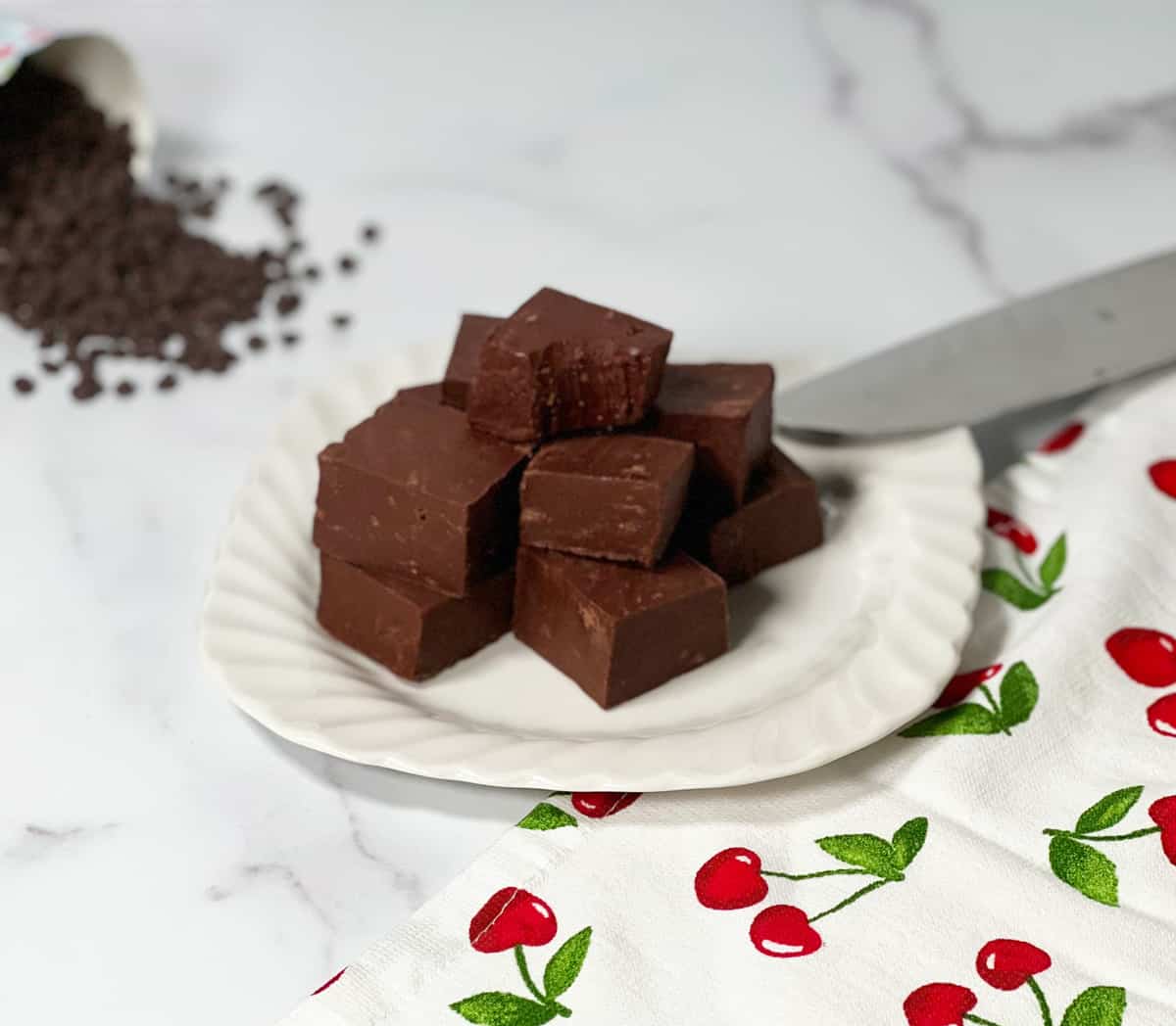 Small white plate with stack of fudge pieces and pretty napkin with cherries on the side and chocolate chips in the background.