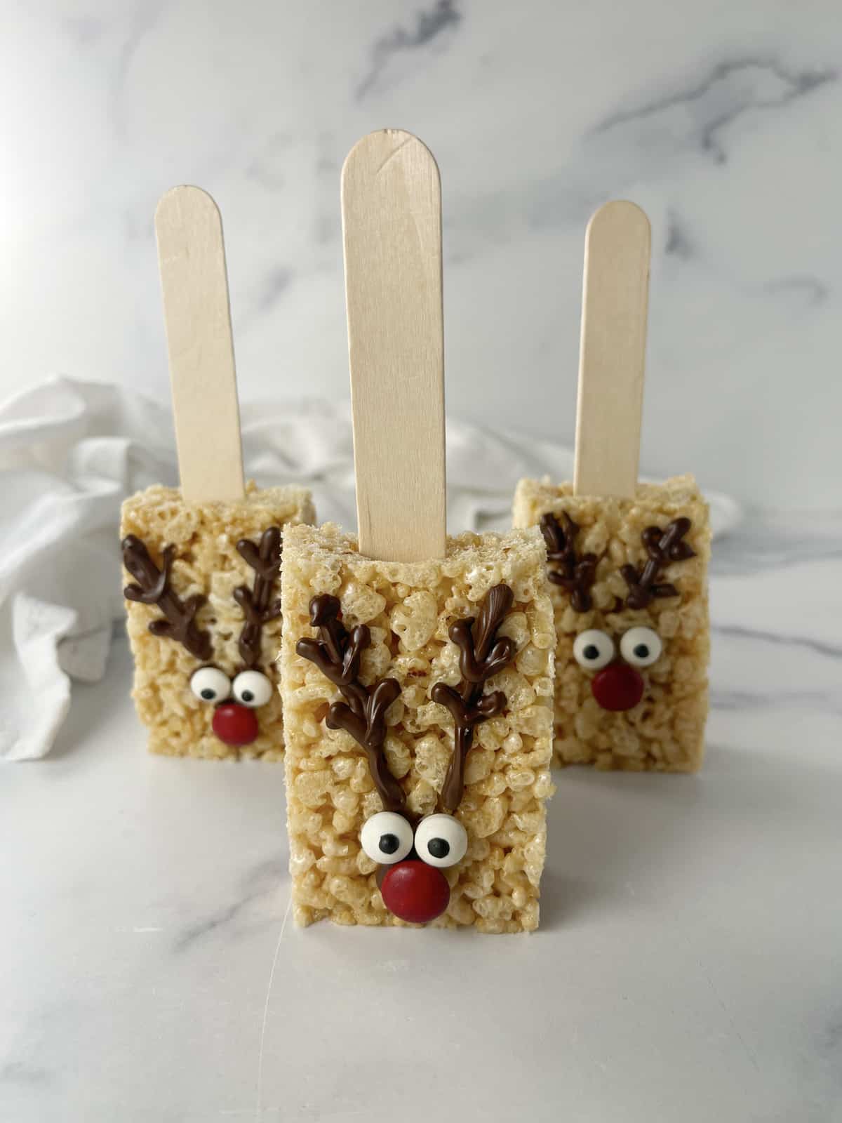 Three rice krispie treats on a popsickle stick with reindeer faces.