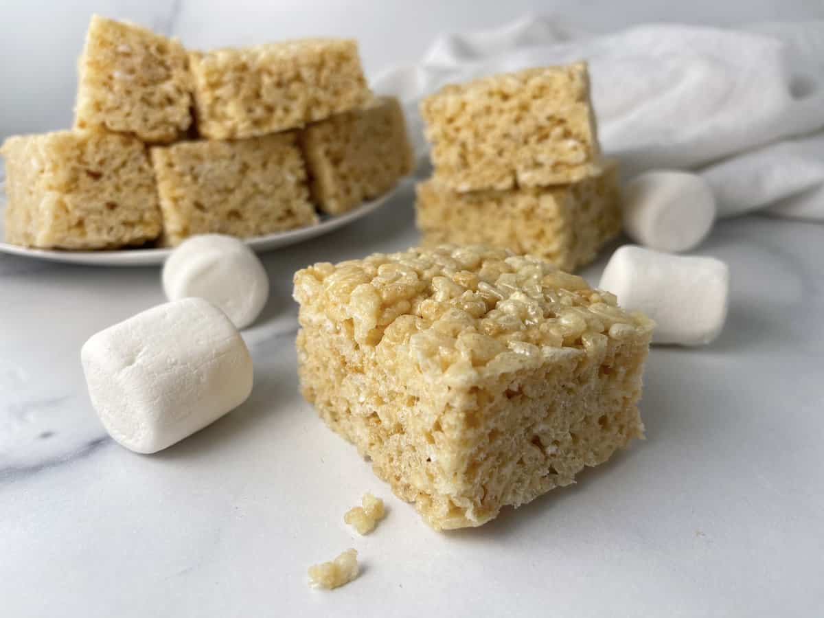 Rice krispie treats on a white plate on the kitchen counter with marshmallows on the side.