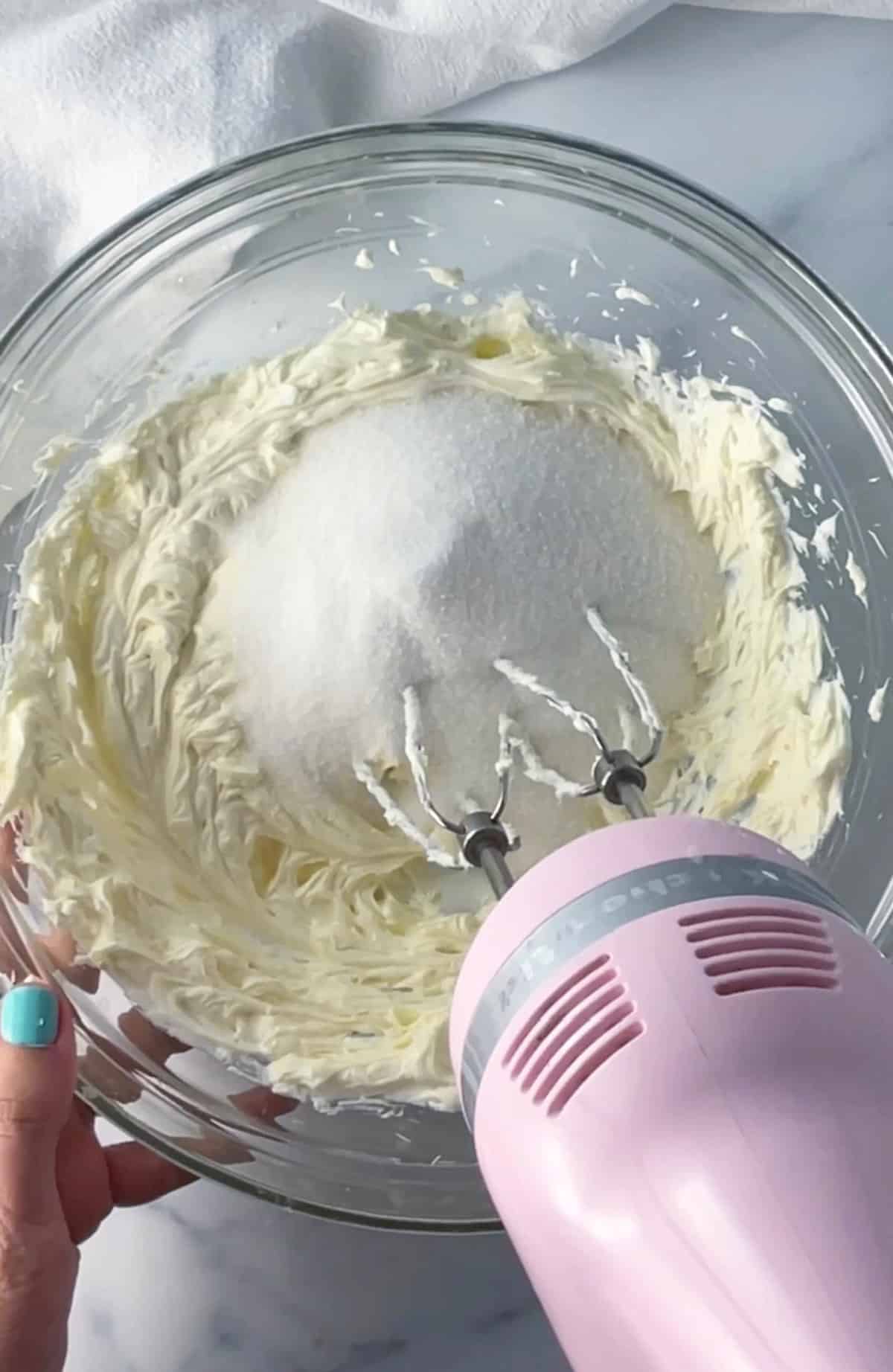 Clear bowl with cheesecake batter being mixed with electric mixer.