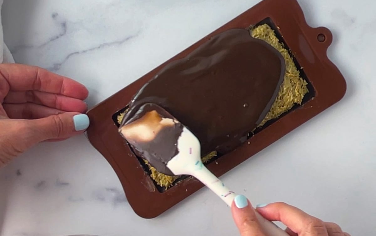 Spreading glossy melted chocolate over pistachio cream and kataifi layer.