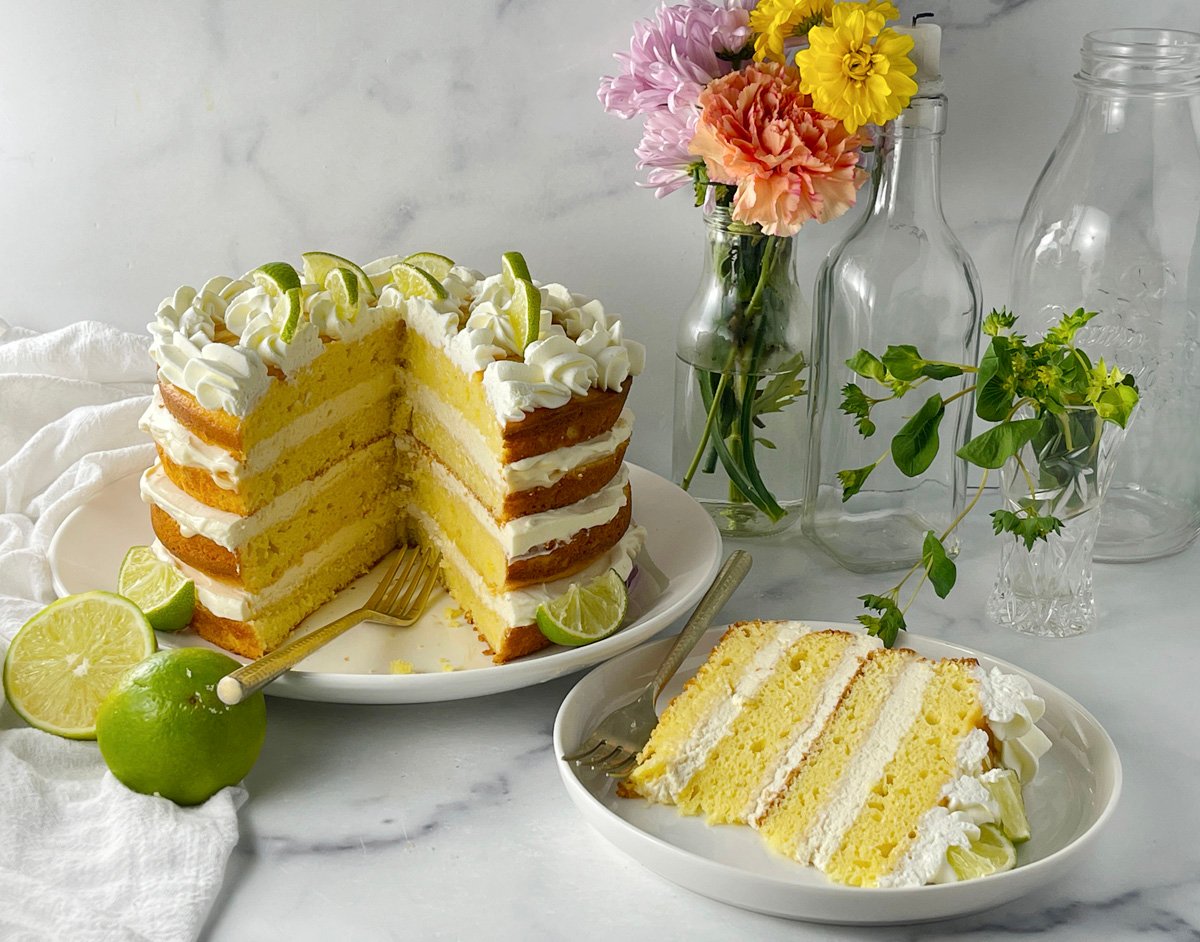 Key lime cake on serving plate with decorative whipped cream border and fresh lime wedges.