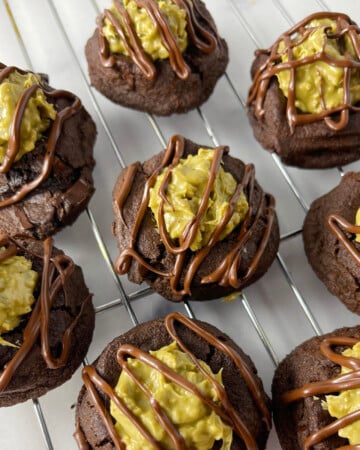 Cooling rack filled with freshly baked Dubai chocolate thumbprint cookies topped with pistachio cream filling and chocolate drizzle.