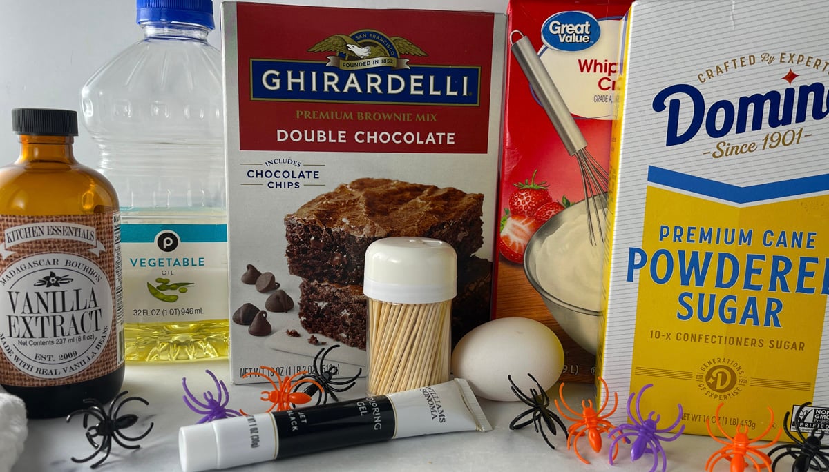 Baking ingredients for Halloween spider web cookie cake arranged on a table, including brownie mix, butter, powdered sugar, cream, vanilla, and black food coloring.
