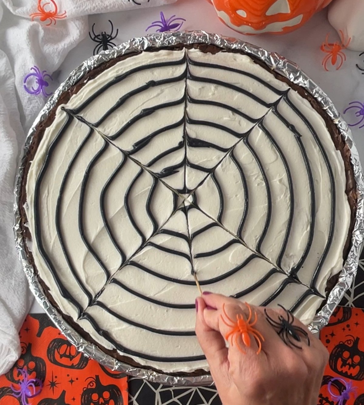Hand using a toothpick to drag lines through black spiral frosting on a cookie cake to create a spider web design.