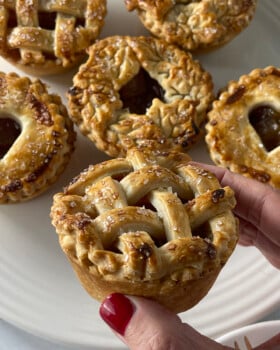 Hand holding a mini caramel apple pie with a golden lattice crust, with more small pies in the background on a white plate.
