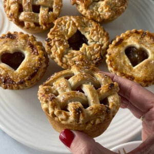 Hand holding a mini caramel apple pie with a golden lattice crust, with more small pies in the background on a white plate.
