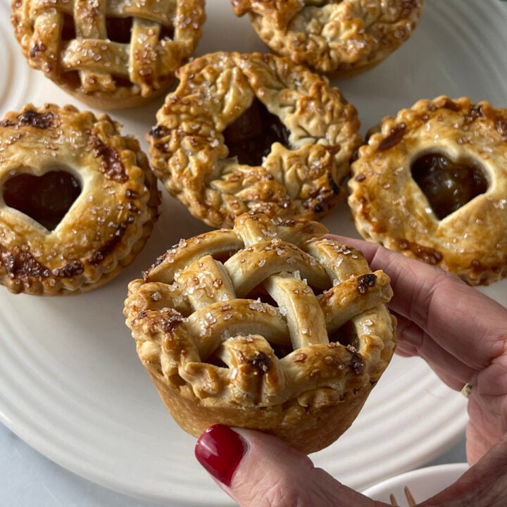 Hand holding a mini caramel apple pie with a golden lattice crust, with more small pies in the background on a white plate.