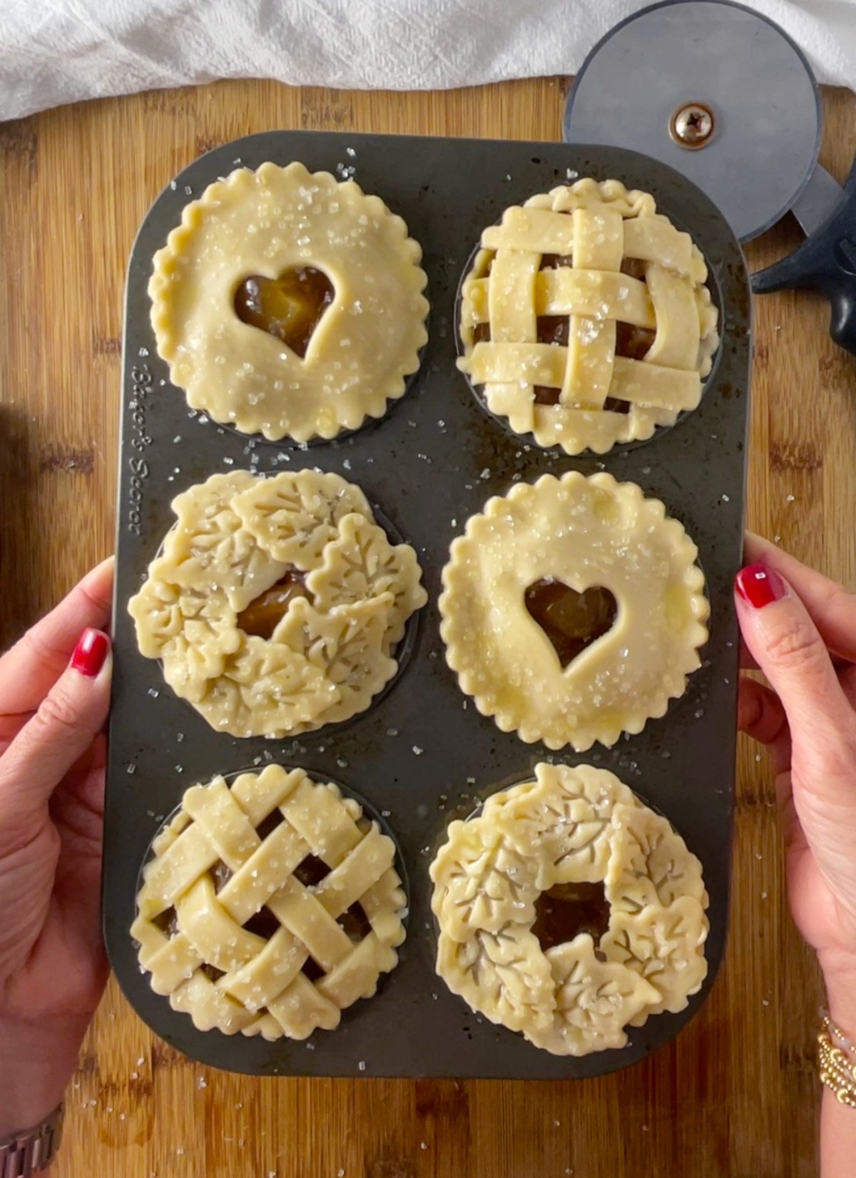 Muffin pan filled with unbaked mini caramel apple pies featuring lattice, heart, and leaf pie crust designs.